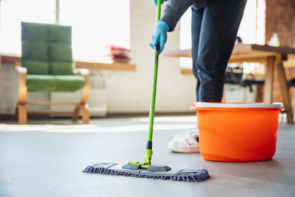 Person mopping a clean floor with a green mop and orange bucket in a bright room.